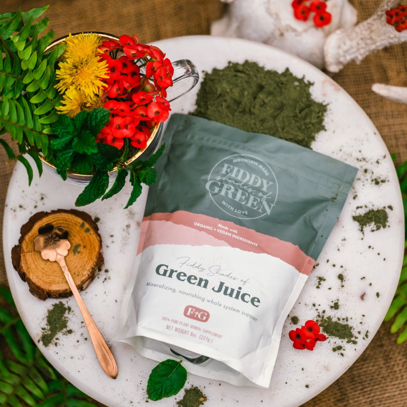 A bag of Fiddy Shades of Green Juice sitting on a marble tray with a glass mug full of bright botanical plants.